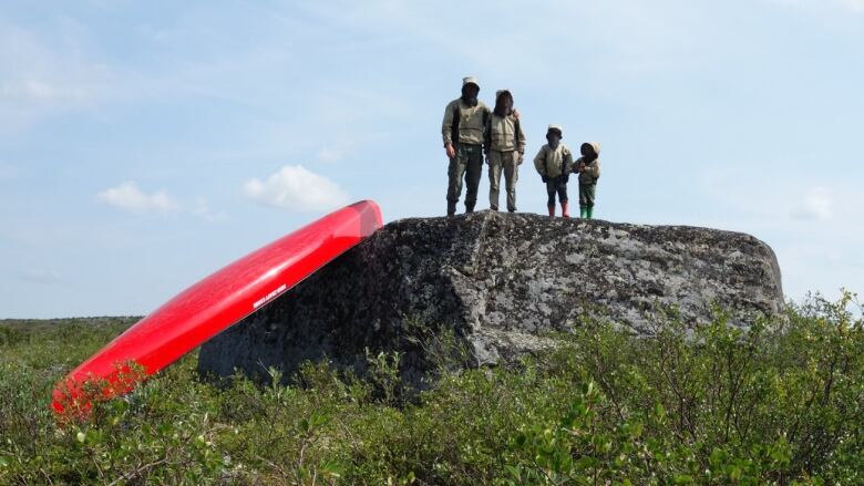 A family stands on a large rock with a canoe resting beside them.