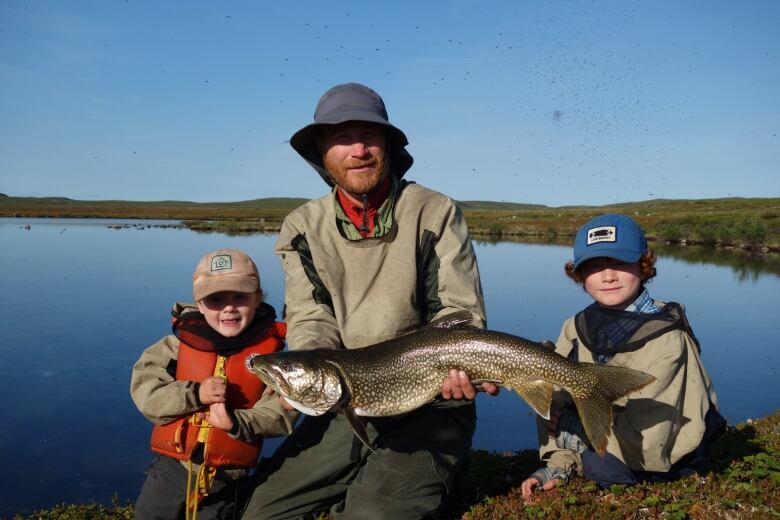 A man kneeling beside a lake with two young boys holds a large fish.