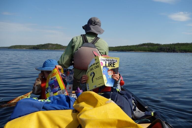 A man sits in the bow of a canoe on a lake with his two young sons sitting behind him, reading magazines.