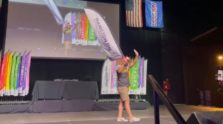 A person standing on a stage holding a banner with the word 'Hamilton.' A North American Gay Amateur Athletic Alliance banner is in the background.