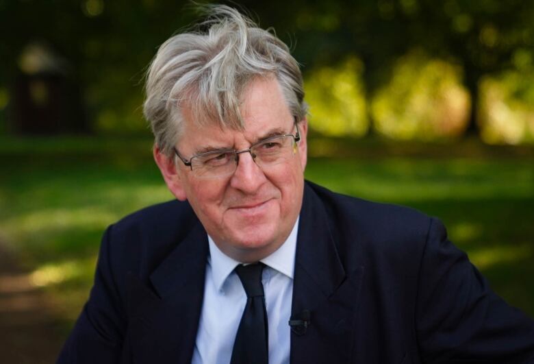 A grey haired man wearing glasses and a blue tie and blazer, sitting on a park bench