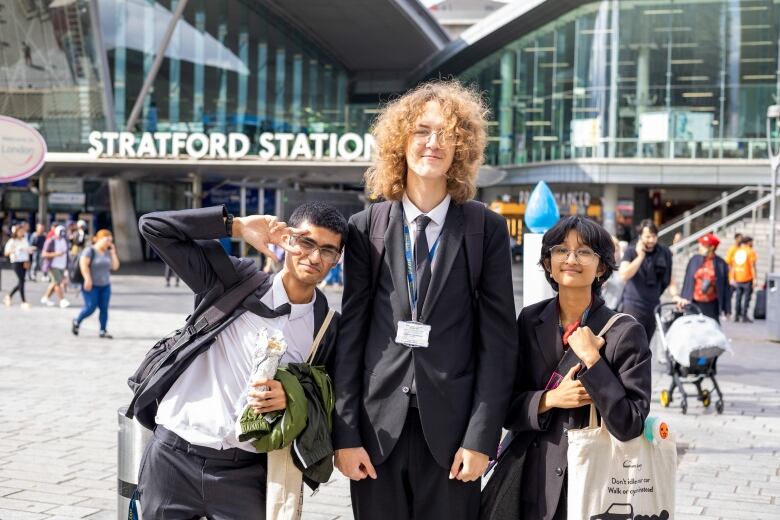 Three students, two boys and a girl, pose for a photo outside a train station.