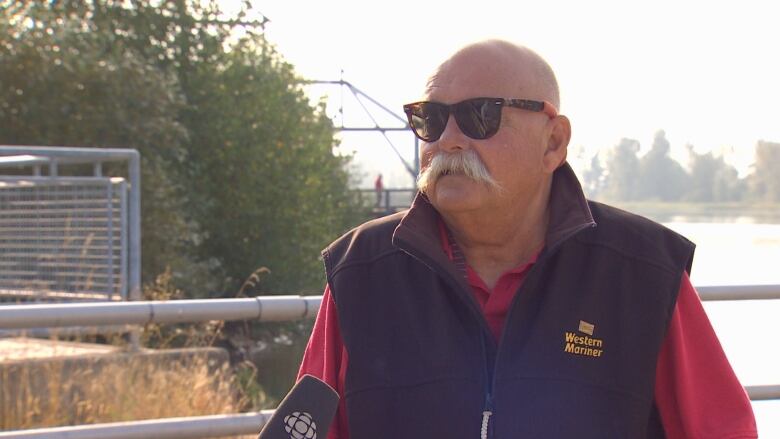 A bald white man with a white moustache looks at the camera, with a body of water behind him.