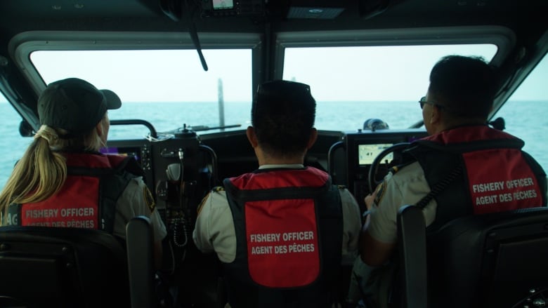 Three people sit at the controls of a boat, with the words 'Fishery Officer' behind them.