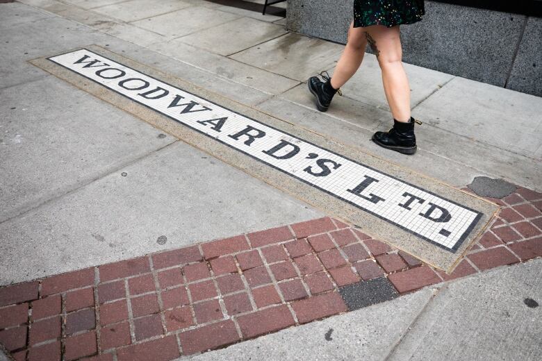 A person walks past a sign on the pavement that reads 'Woodward's Ltd.' in old time font.