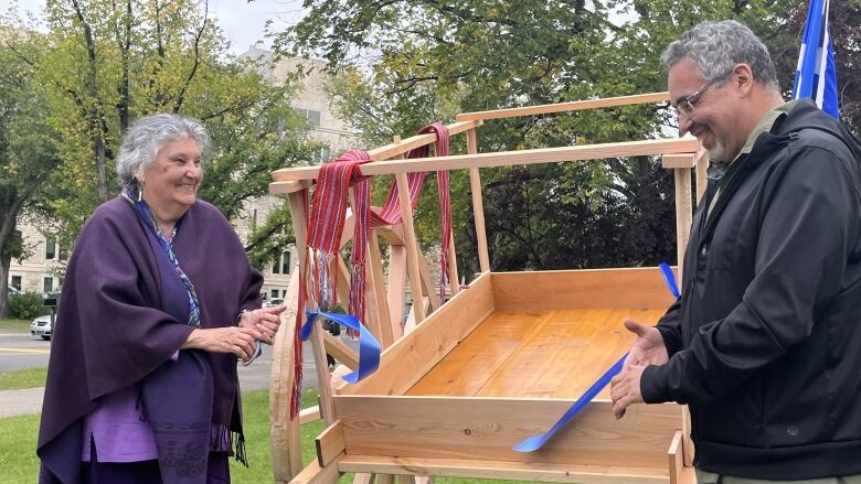 Two people stand beside a wooden cart, with a blue ribbon tied to it and cut in the middle