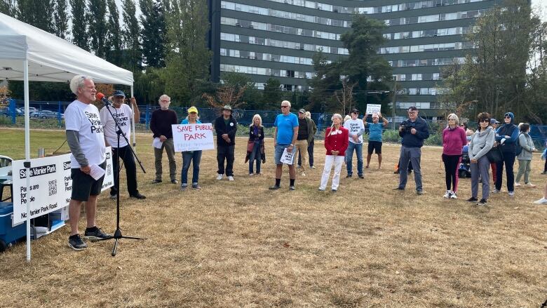 A group of old people gather around a tent in a park. One of them is holding a mic.