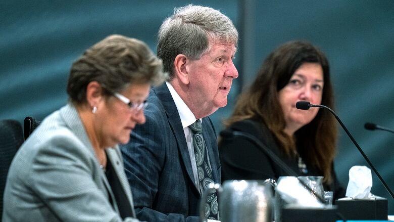 Three white people, a man and two women, sit at a long table with microphones