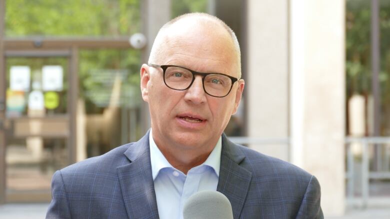 A man wearing glasses and a suit is speaking into a microphone, with a building in the background.