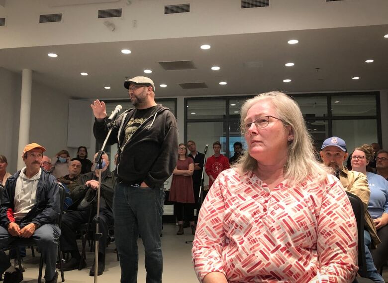 A woman with white hair and glasses listens intently as a man speaks at a microphone.