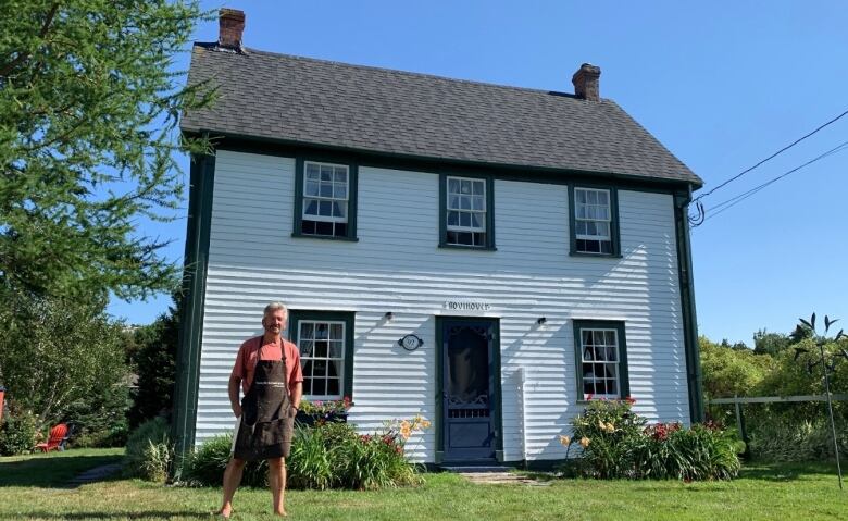 A man in an apron stands in front of a white two-storey house with dark trim.