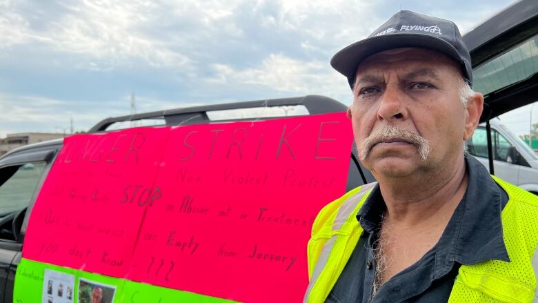 A man with a cap stands beside a sign taped to a car that says Hunger Strike. 