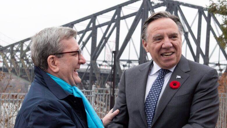 Two men smile in front of a bridge.