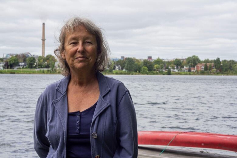 A woman stands by the water, with a chimney in the background.