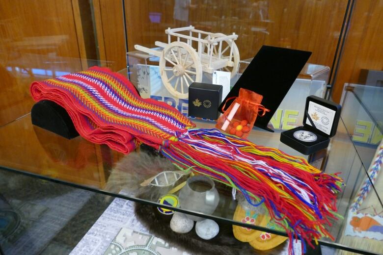 A multi-coloured sash, a beaded flower, and a minature wooden cart are displayed inside a glass case.