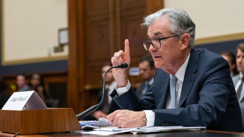 A man with white hair and glasses, and wearing a blue suit, points his right index finger in the air as he sits at a desk and speaks into a microphone.