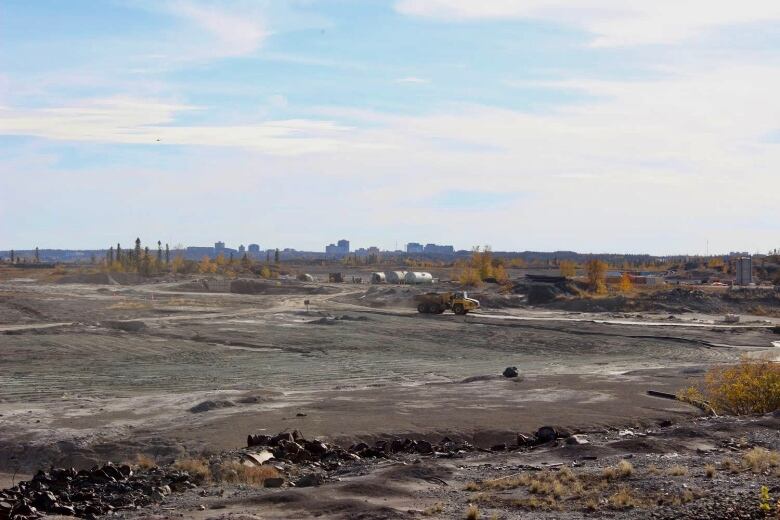 A wide photo with a dump truck rolling over a dusty road and a city skyline in the distance.