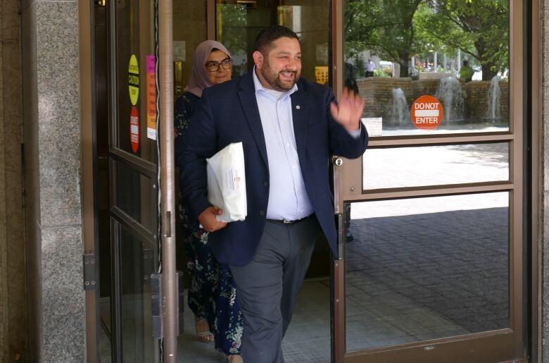 A man in a suit holds a large white envelope under his arm, waving and smiling as he walks out the door of a public building.