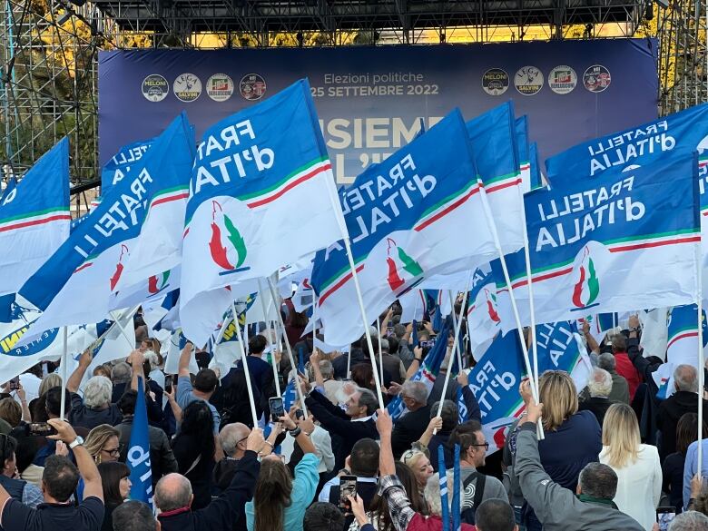 A large group of people hold flags with italian writing on them, in front of an election sign.