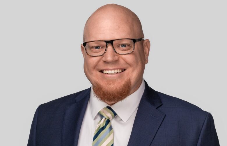 A bald man with a red beard and glasses smiles while wearing a suit.