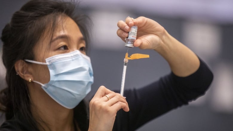 A woman with long black hear who is wearing a mask holds a syringe.