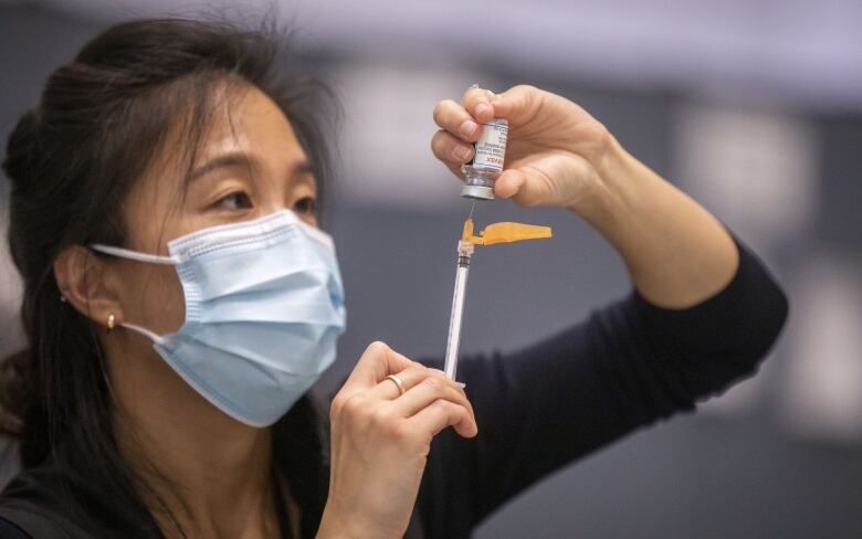 A woman with long black hear who is wearing a mask holds a syringe.