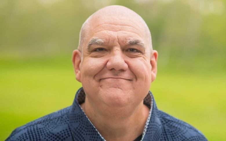 A bald man in a blue collared shirt smiles in front of a green outdoor background, which is blurred.