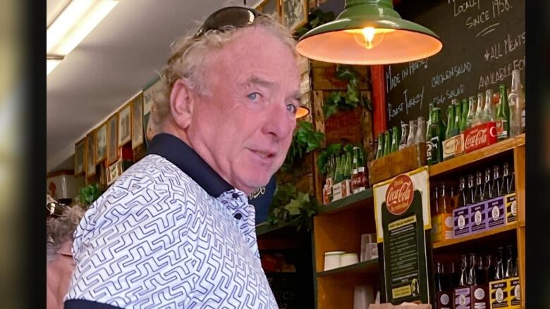 A man in a short-sleeved shirt stands at the counter of a restaurant, with a chalkboard and shelves lined with pop bottles behind the counter.