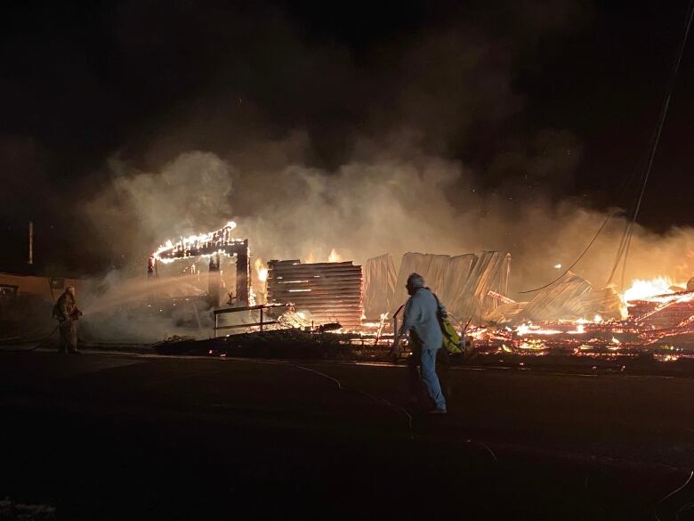 A building in a dark night completely engulfed in flames, with a few people walking nearby.