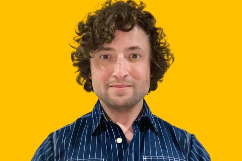 A man with curly hair wearing a striped blue long sleeve shirt smiles in front of a vibrant orange background.