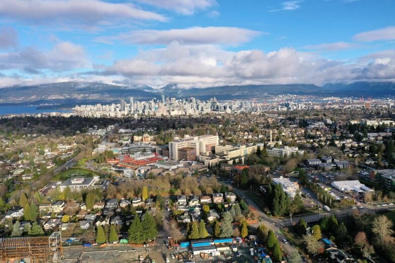 An aerial photo shows a hospital campus surrounded by trees and single-family homes, with the Vancouver skyline, English Bay and the North Shore mountains in the background.