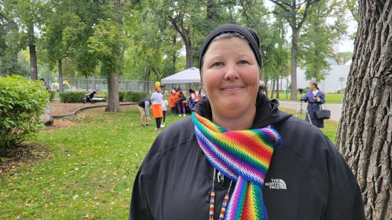 A woman wearing a black jacket and a rainbow sash scarf smiles in a park. Behind her, people wearing orange are set up below a canopy.