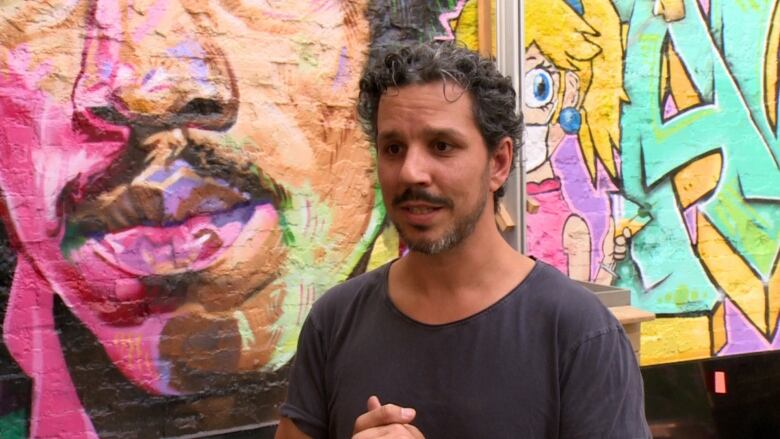 A bearded man clasps his hands in front of a mural wall.