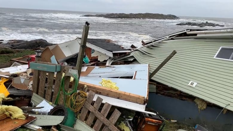 The wreckage of a home lies strewn against the surf. A wall lies on top of the pile, having been peeled from its structure like the skin of an orange.