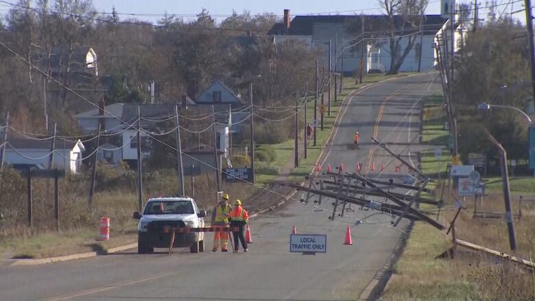 Several power poles lie in a road after being snapped off at the base. Crews stand by a barrier preventing traffic from going down the street.