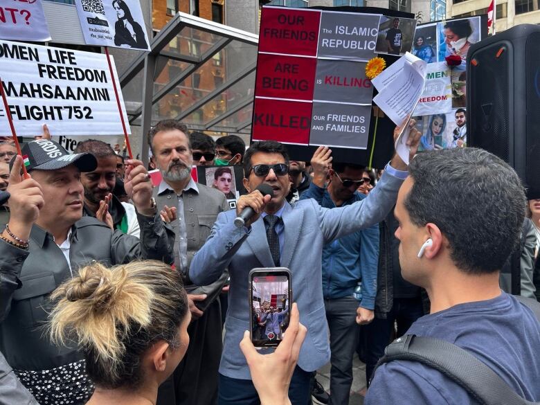 A man in a suit and sunglasses speaks into a microphone while protesters holding signs gather around him and film him.