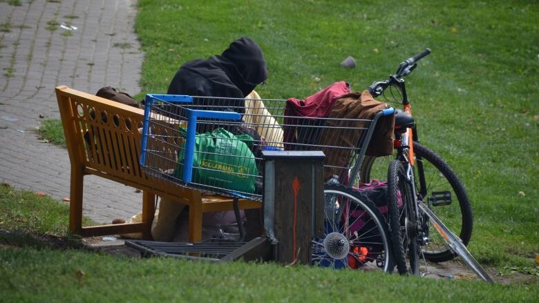 A man sits on a bench in a park surrounded by his possessions, including a shopping cart and a bicycle. 