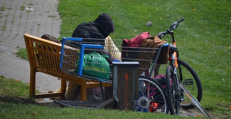 A man sits on a bench in a park surrounded by his possessions, including a shopping cart and a bicycle. 