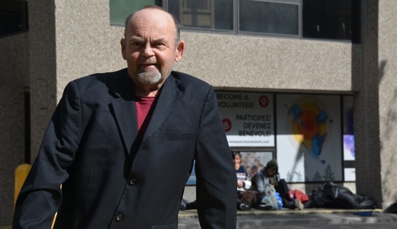 Bob Johnston stands in Memorial Park in downtown Sudbury wearing a suit jacket, with some people sitting on the ground behind him. 