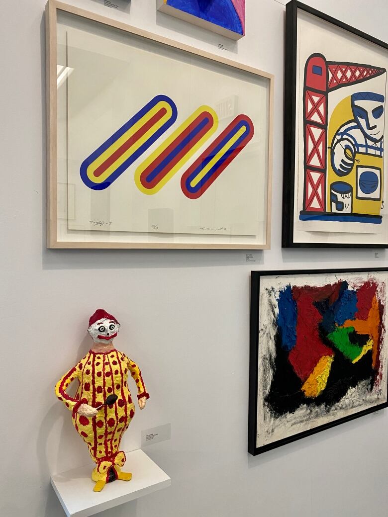 A sculpture of a clown and prints of abstract art seen in an exhibit. 