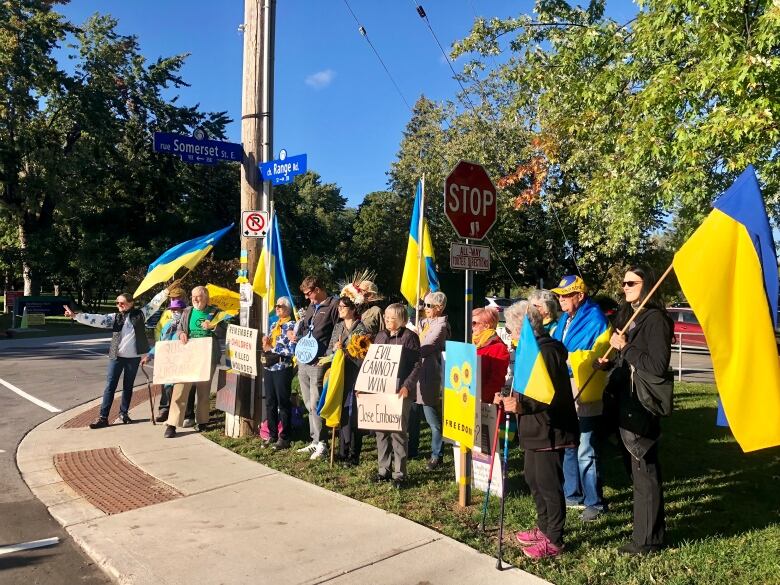 A group of people waving Ukrainian flags stands beside a road on a sunny day.