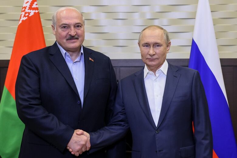 Two men, both wearing blue suit jackets, shake hands in front of the flags of Russia and Belarus. 