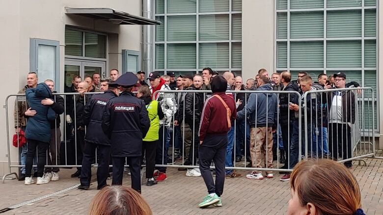 A group of men stand in a holding pen of sorts outside of a building as guards look on. 