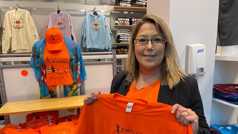 A woman wearing glasses holds an orange t-shirt that says Every Child Matters. Behind her are stacks of more orange t-shirts.