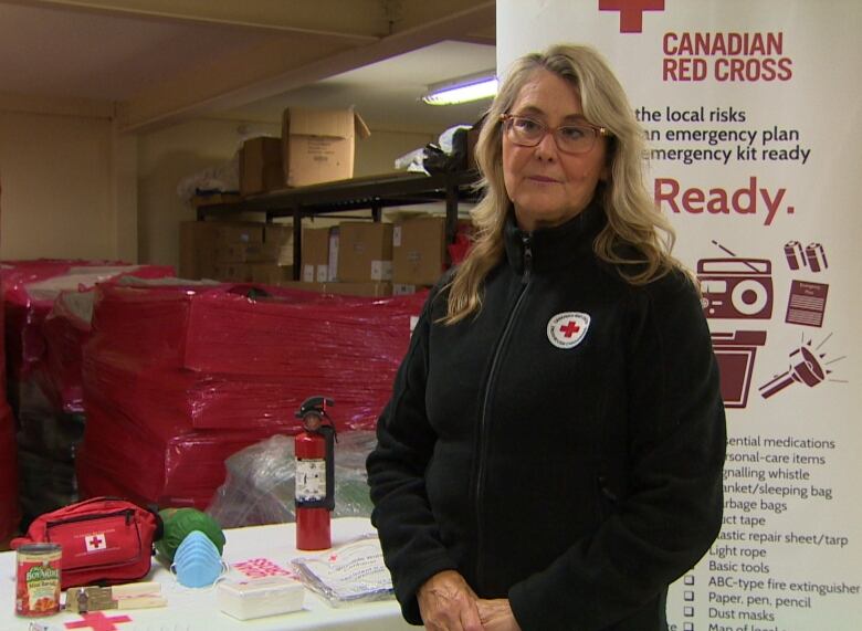 A person with long hair and glasses wearing a black fleece stands next to a table carrying emergency supplies like a first aid kit and a fire extinguisher.