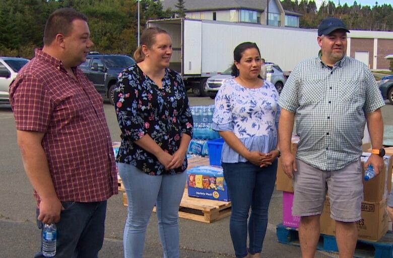 Four people stand in a parking lot. Pallets of supplies, including water and potato chips, can be seen behind them.