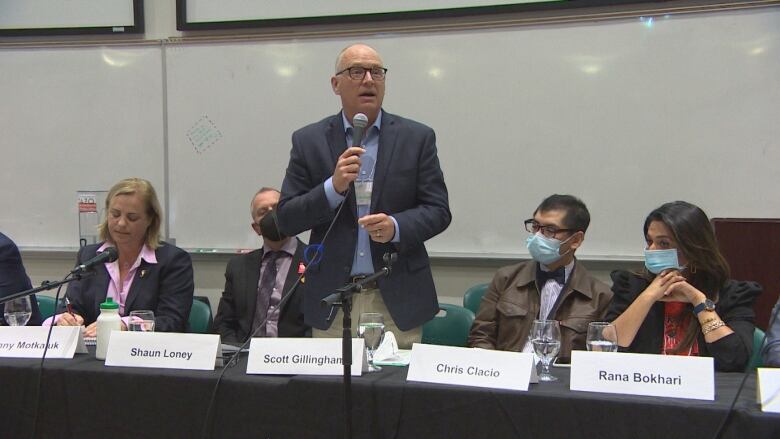 A man in a suit is standing and speaking into a microphone, while other people sit at a table on either side of him. 