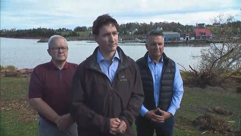 Prime Minister Justin Trudeau, wearing a windbreaker, stands in front of a river with a local Member of Parliament on each side of him. 