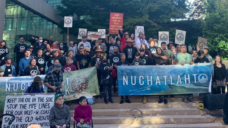 A group of people, many of whom are Indigenous, stand protesting on a series of steps. Many of them are holding up hoardings reading 'Nuchatlaht'.