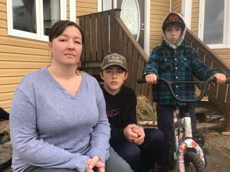 A photo of three people sitting in front of a yellow house. On the left, a person in a purple shirt folds her hand. In the middle, a young teen in a baseball hat folds his had. On the right, a boy sits on a bike.